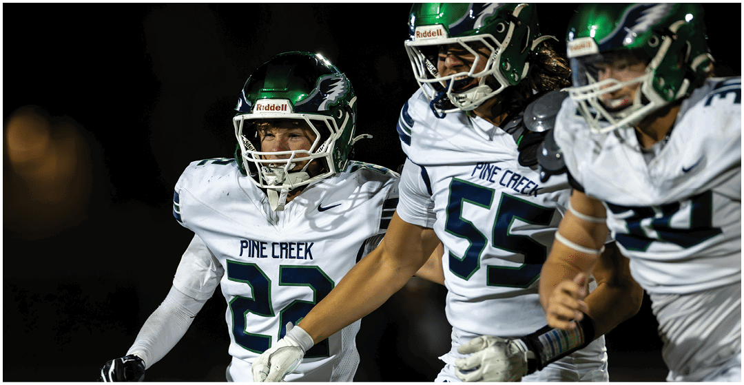 Three people wearing white football uniforms with green helmets and “Pine Creek" on their jerseys are walking together on a field at night. Description generated by AI