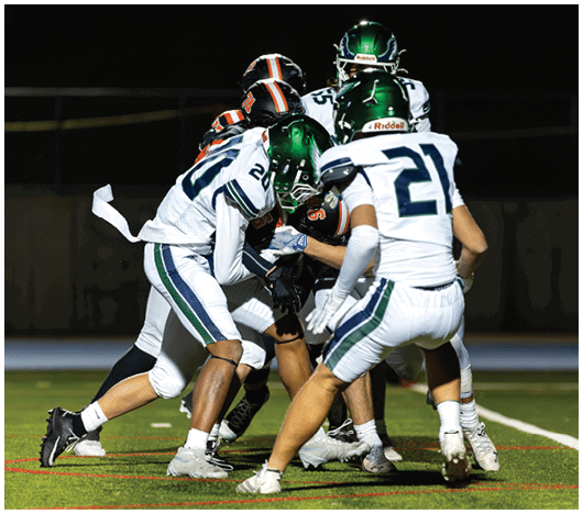 Four football players in white and green uniforms are tackling a player in a black and orange uniform on a field at night. Description generated by AI
