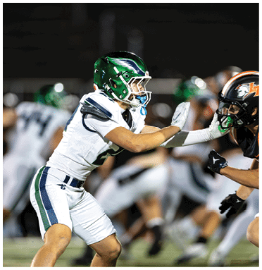 Two people in football uniforms and helmets are engaged in a play on a field, with one person extending their arms toward the other. Description generated by AI