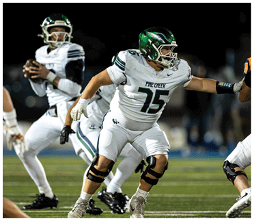 Two people in white football uniforms with green helmets are on a football field, with one preparing to throw a ball and the other blocking an opponent. Description generated by AI