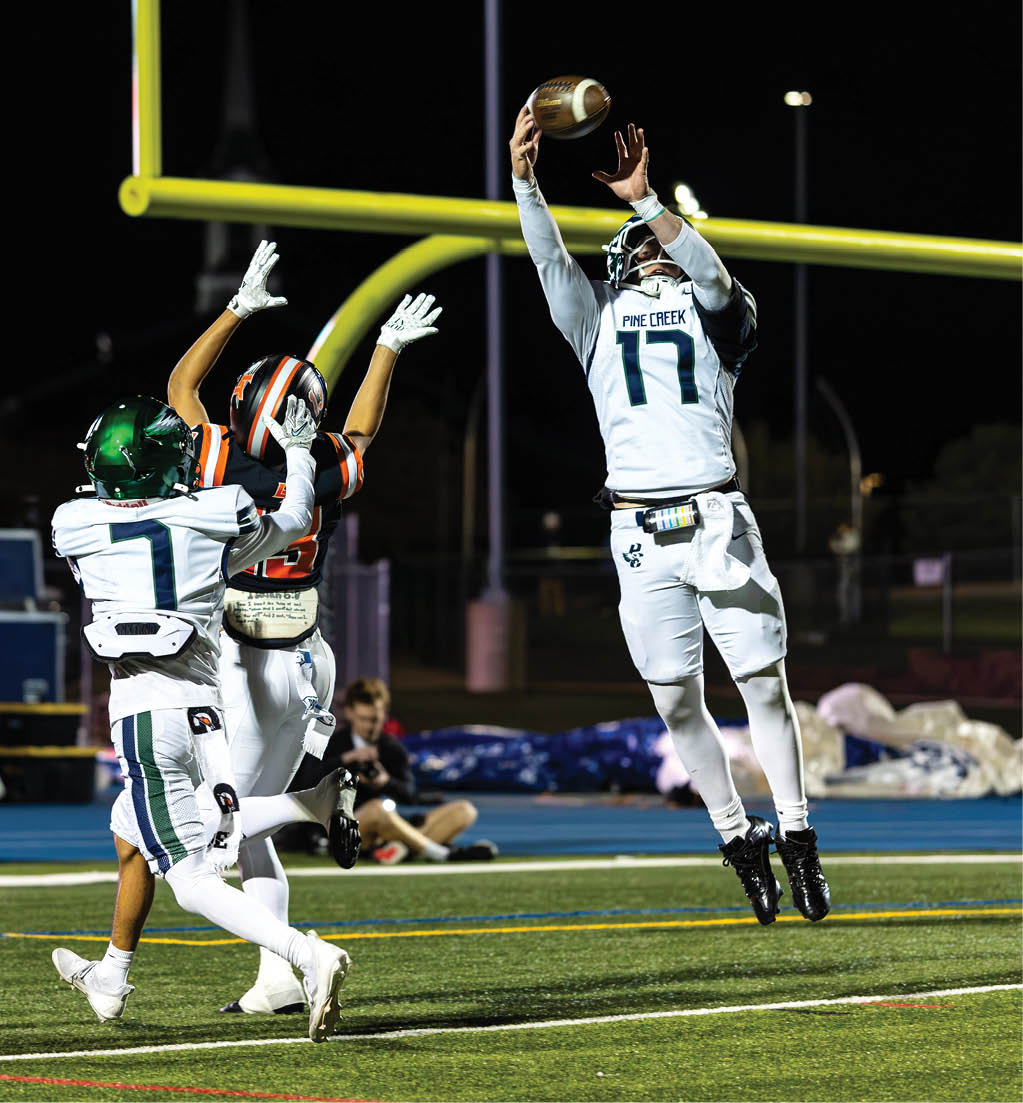 A person in a white football uniform with the number 17 is jumping to catch a football near the goalpost while two opposing players in black and orange uniforms attempt to block the catch on a nighttime field. Description generated by AI