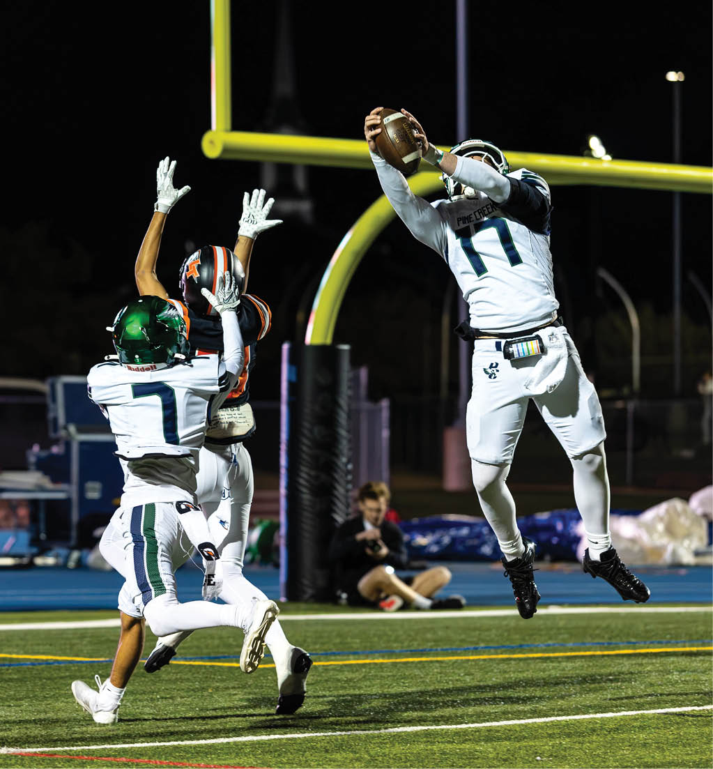 Three football players in action near the goalpost with one player jumping to catch the ball while two others attempt to block or intercept. Description generated by AI