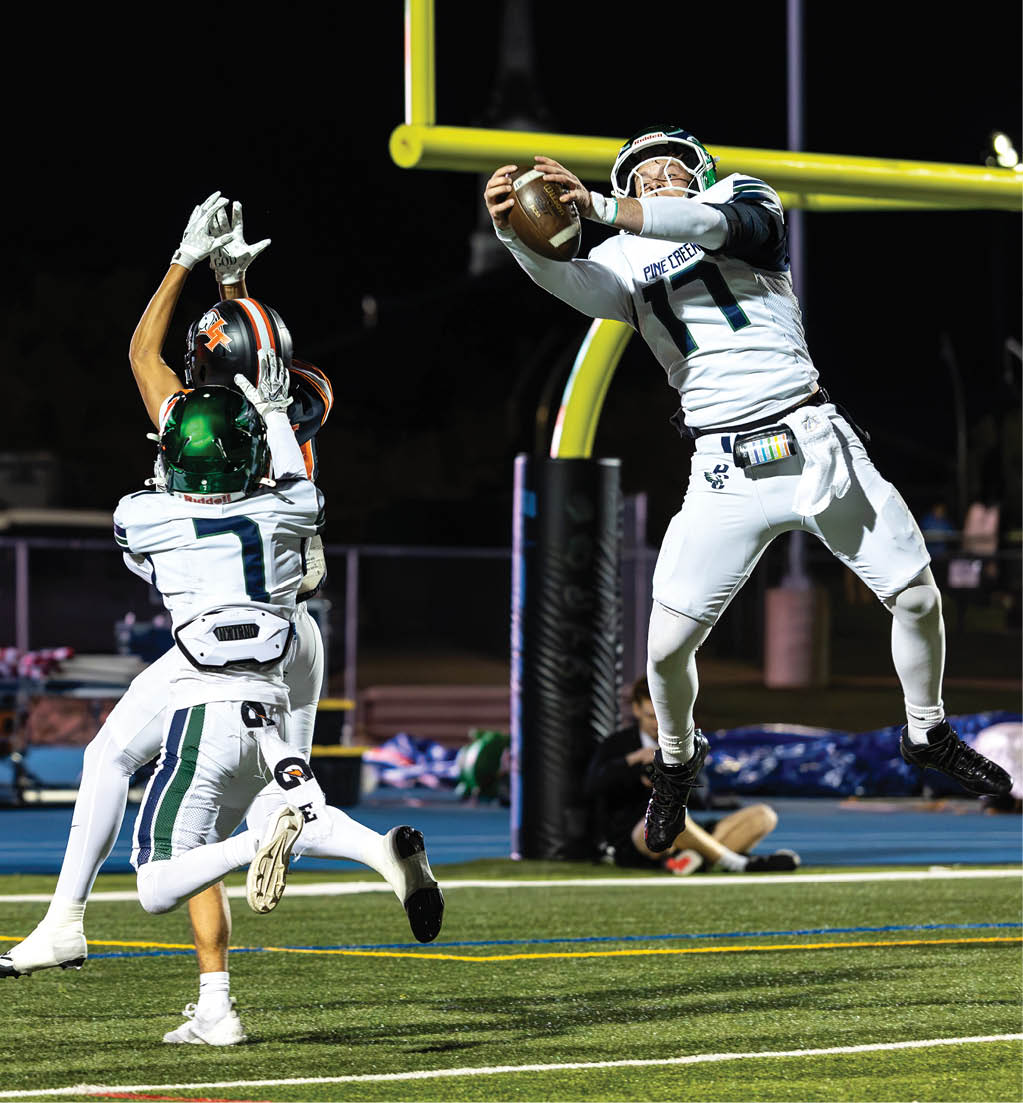 Three people in football uniforms are actively reaching for a football near the goalpost on a field during a night game. Description generated by AI