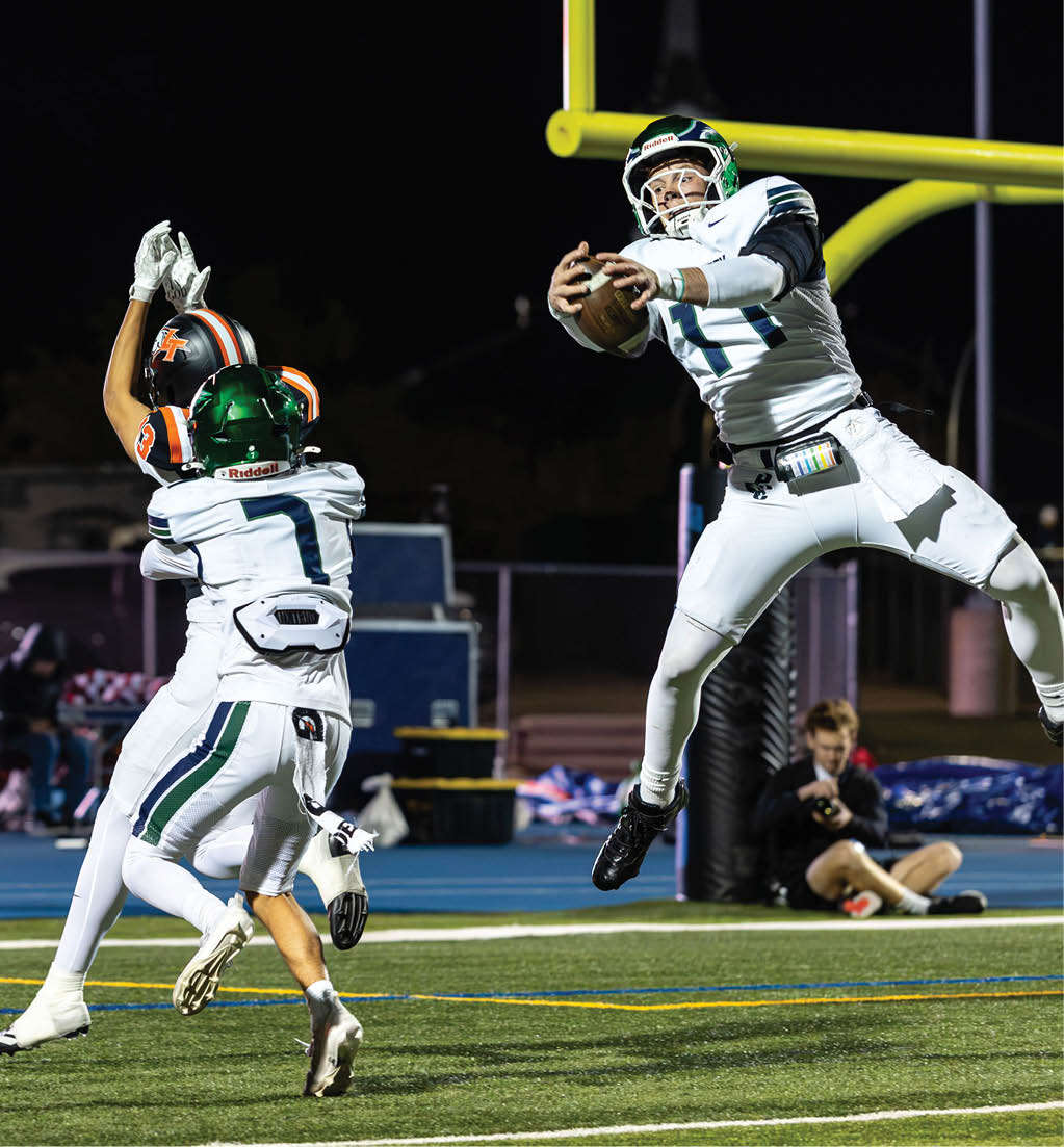 Three people in football uniforms are actively competing near a goalpost on a field at night, with one person jumping to catch the ball while another attempts to block. Description generated by AI