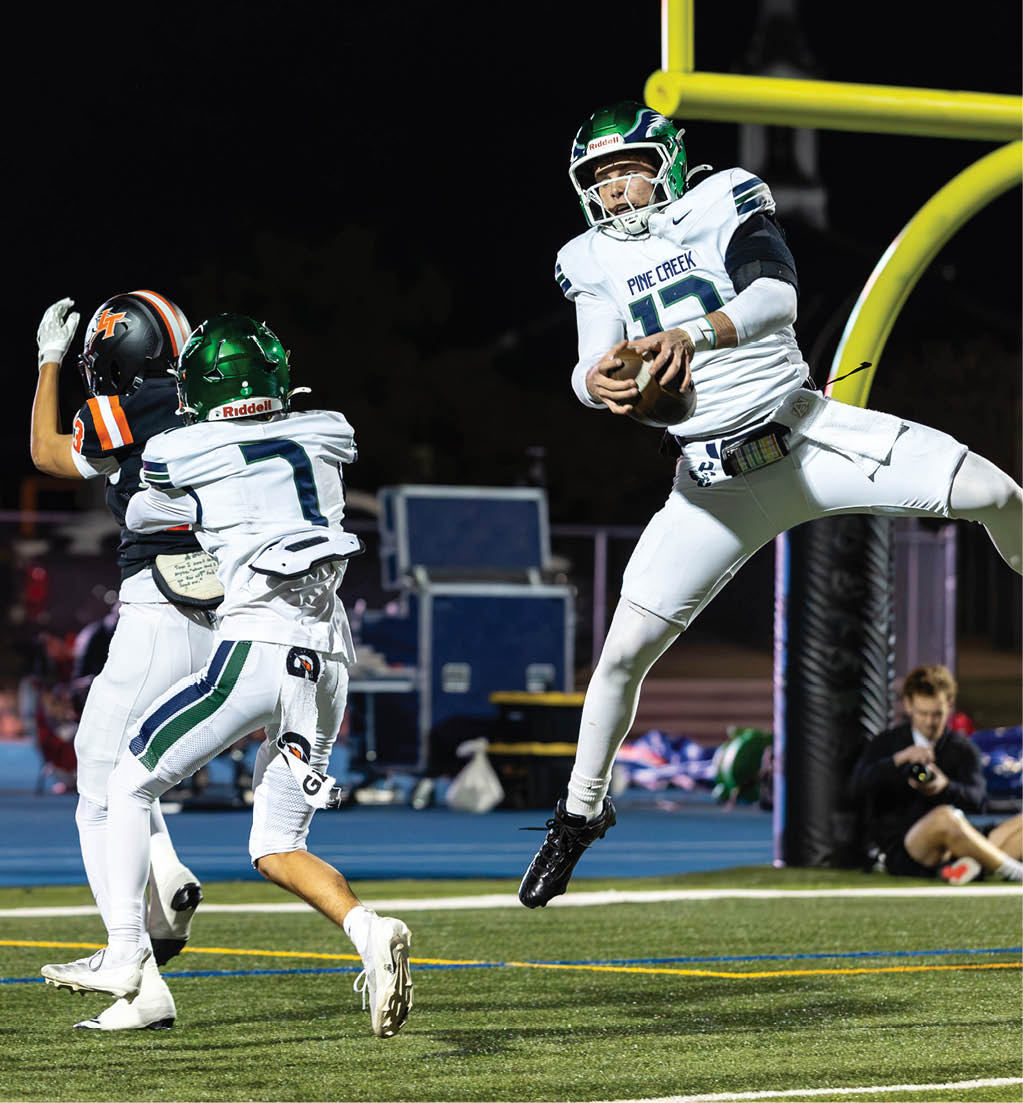 A person in a white football uniform with a green helmet is jumping while holding a football near the goalpost as two other players in white and one player in a black and orange uniform are nearby on a football field at night. Description generated by AI