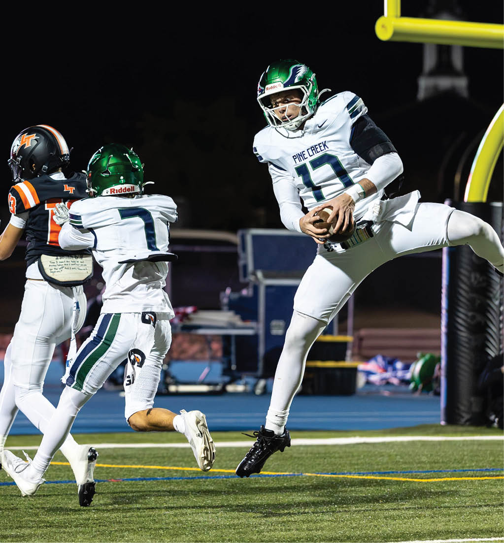 A person in a white football uniform with a green helmet is holding a football and jumping near the goalpost while two other players in white and one player in a black and orange uniform are running nearby on a football field at night. Description generated by AI