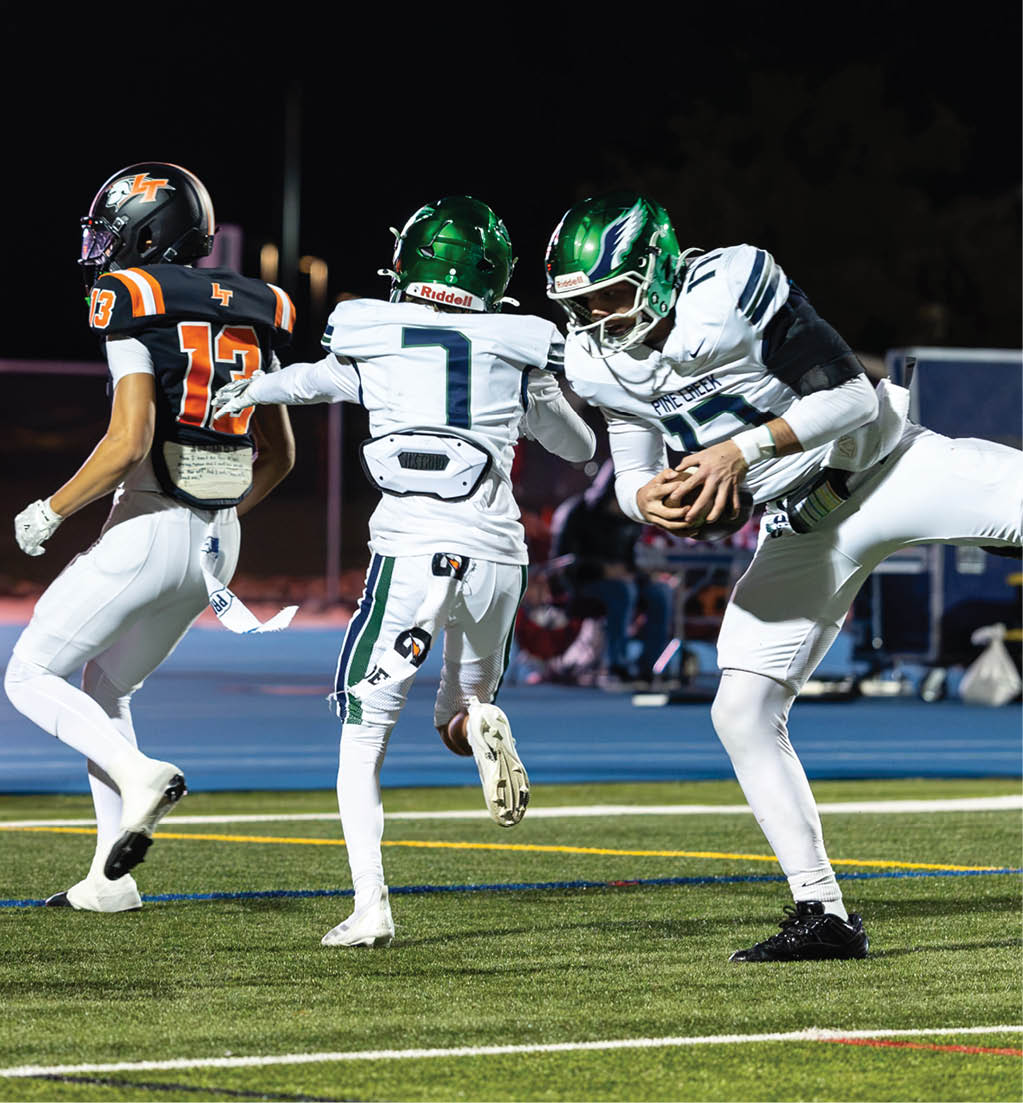 Three people in football uniforms are on a field at night with one person holding a football while another reaches toward a third person running away. Description generated by AI