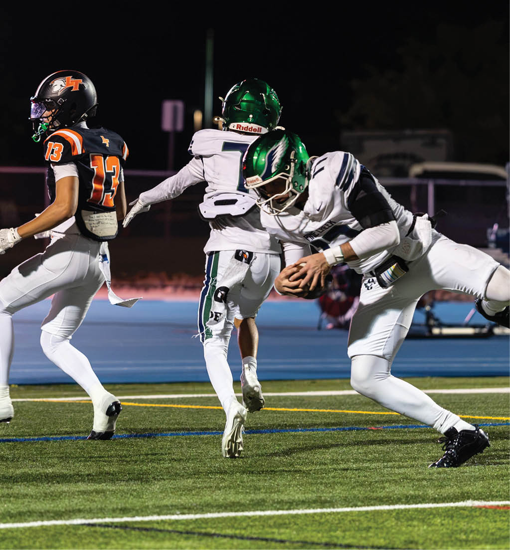 Three people in football uniforms are actively playing on a field at night, with one person in white and green gear holding the ball while another in black and orange runs nearby. Description generated by AI