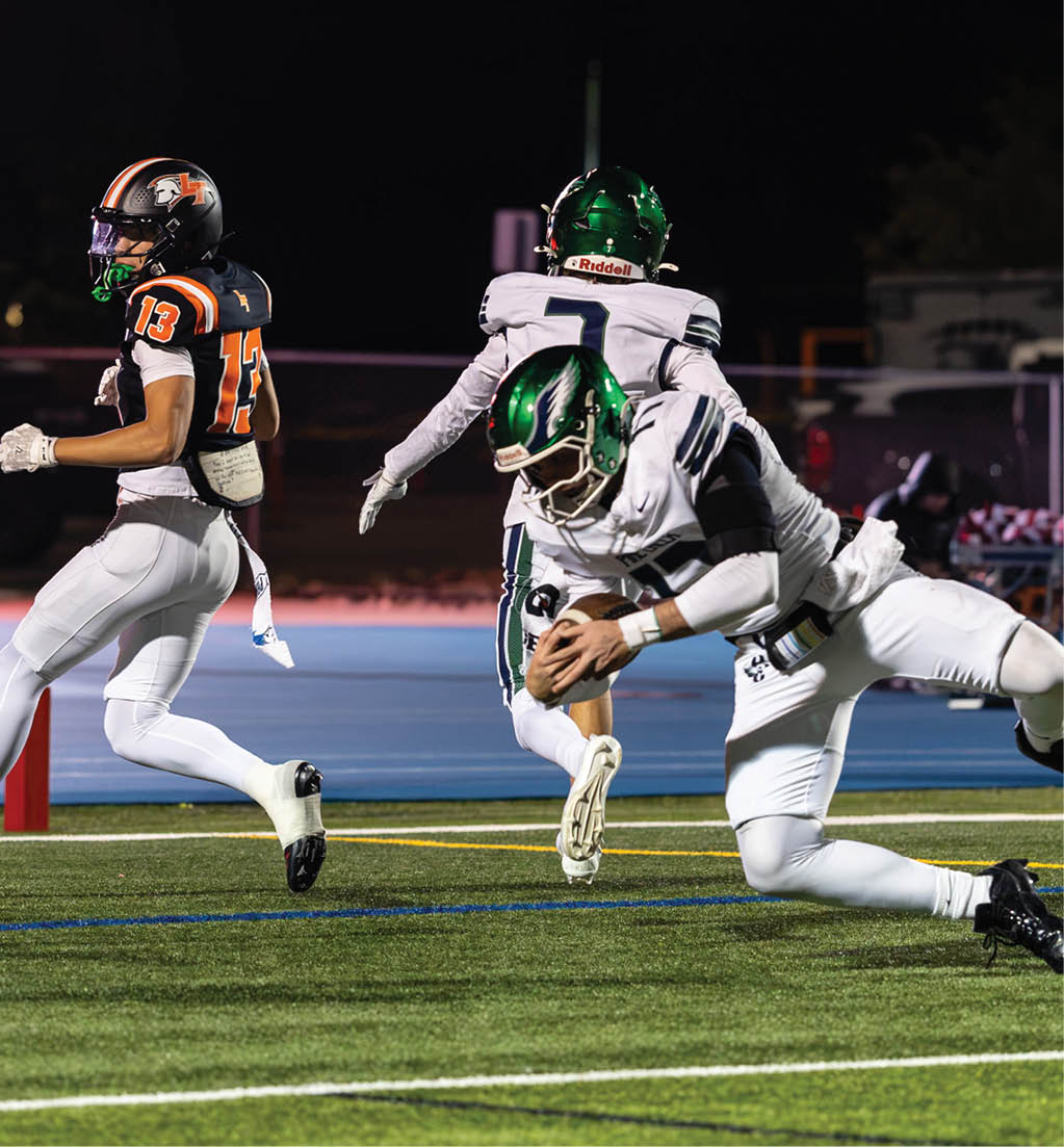 A person in a black and orange football uniform is running while two people in white and green football uniforms are tackling or chasing each other on a football field at night. Description generated by AI