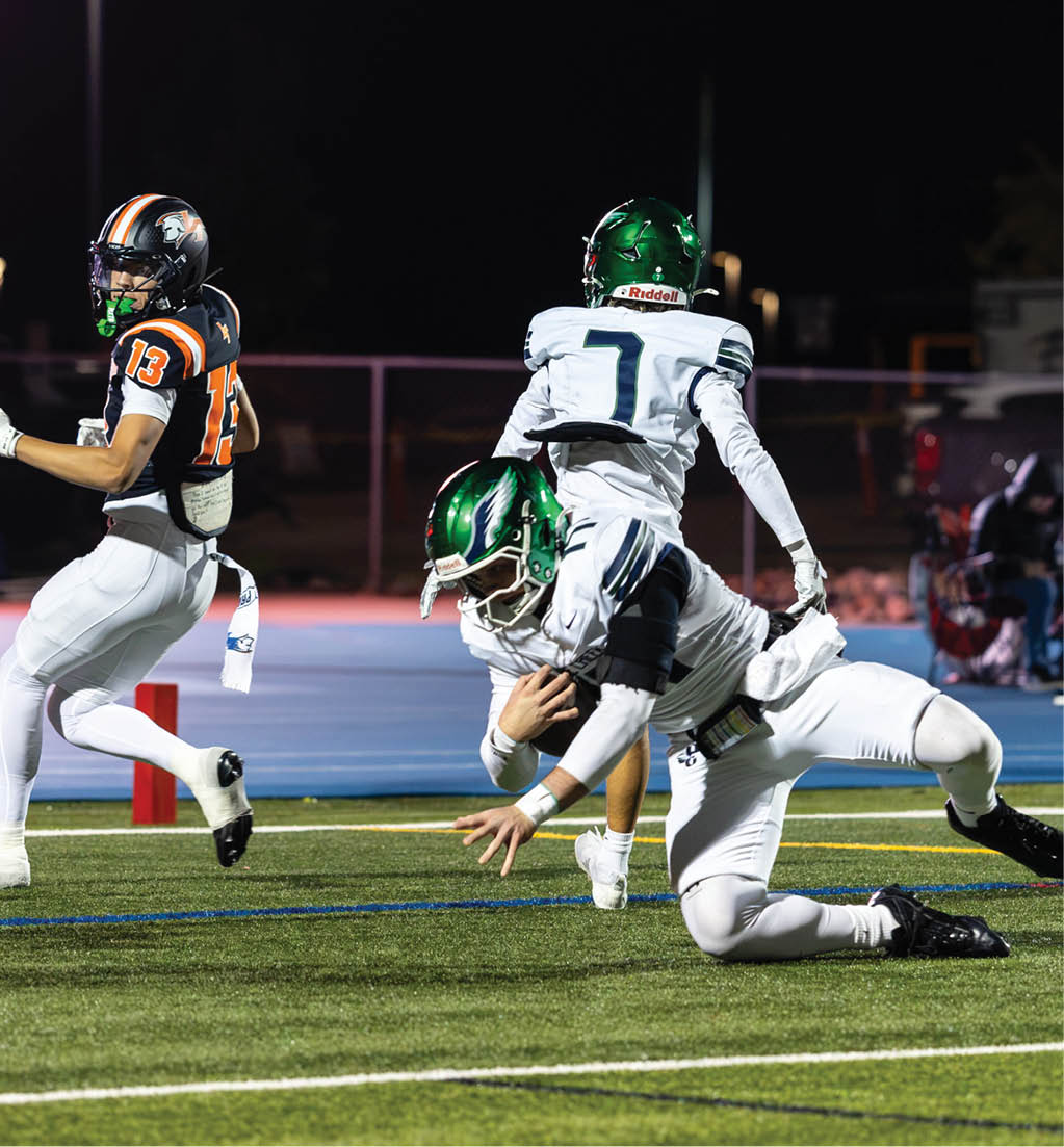 Two people in white football uniforms with green helmets are tackling a person in a black and orange football uniform on a field at night. Description generated by AI