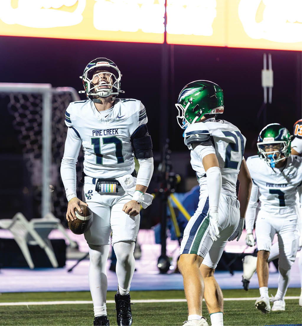 Three football players in white uniforms with green helmets are on a field, with one player holding a football and looking upward. Description generated by AI