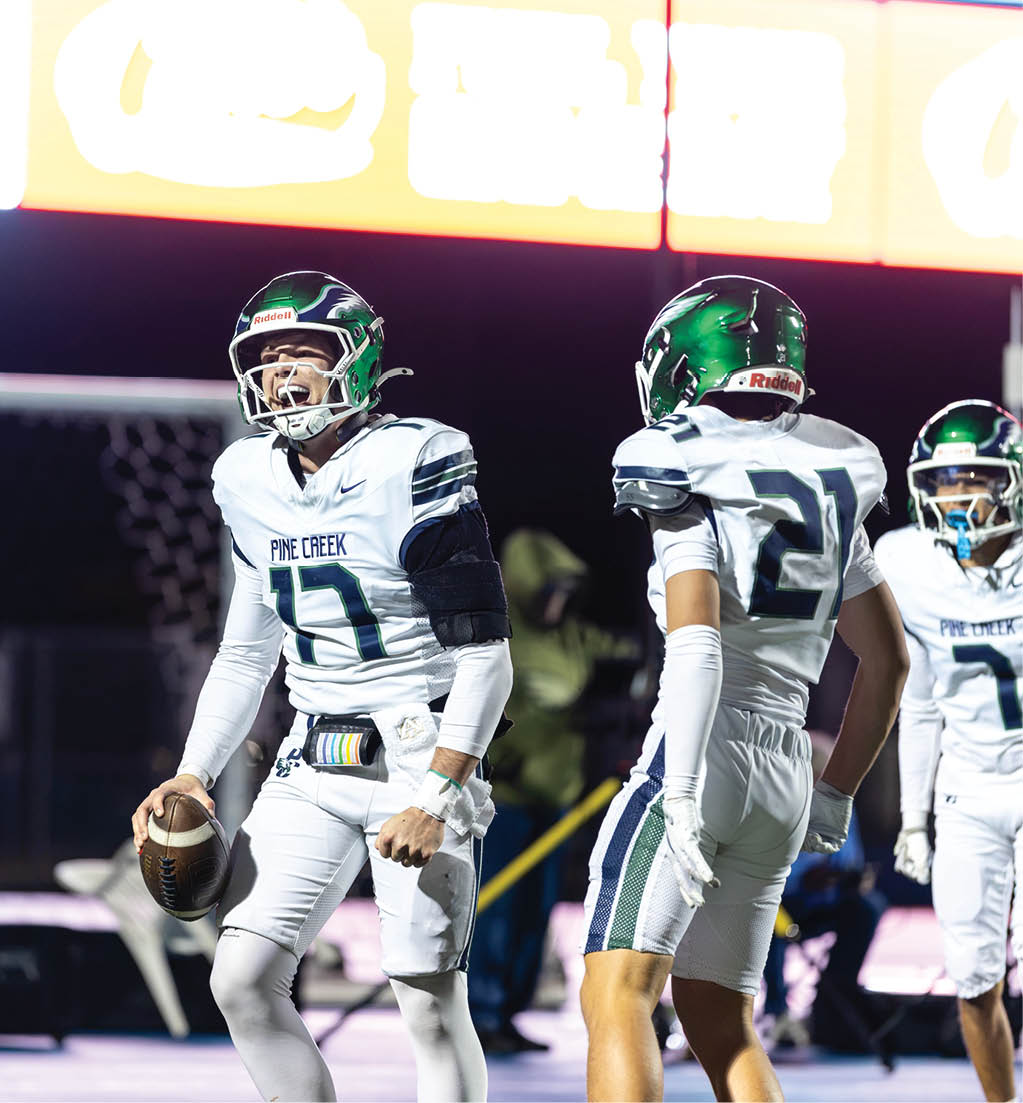 Three people in football uniforms with green helmets are on a field, with one holding a football and appearing to shout or communicate. Description generated by AI