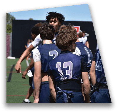 A person in football gear and helmet is crouched on a field with other players lined up in the background during a game. Description generated by AI