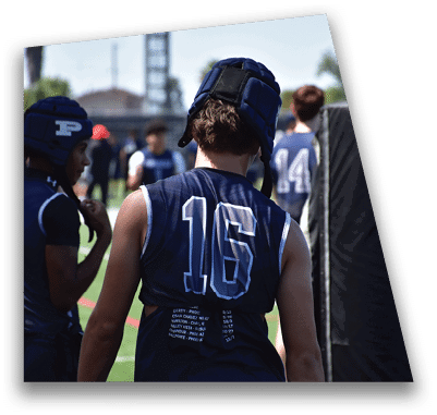 A person in football gear and helmet is crouched on a field with other players lined up in the background during a game. Description generated by AI
