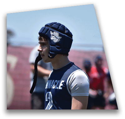 A person in football gear and helmet is crouched on a field with other players lined up in the background during a game. Description generated by AI