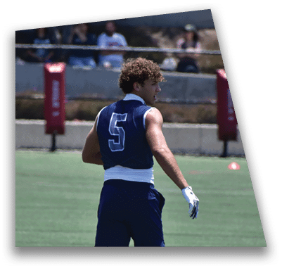 A person in football gear and helmet is crouched on a field with other players lined up in the background during a game. Description generated by AI