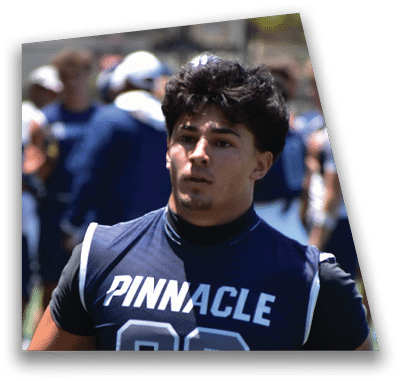 A person in football gear and helmet is crouched on a field with other players lined up in the background during a game. Description generated by AI
