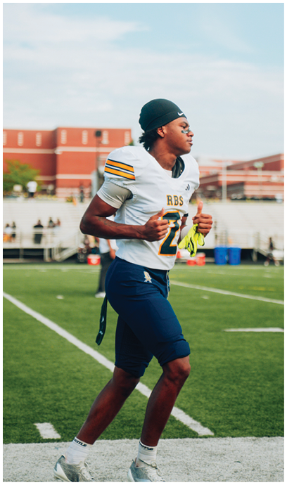 A person wearing a football uniform with a helmet in hand is jogging on a green field near the sideline with a school building and bleachers in the background. Description generated by AI