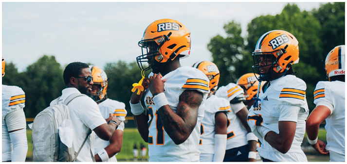 Adults wearing orange helmets and white football uniforms are gathered on a field with a coach giving instructions. Description generated by AI