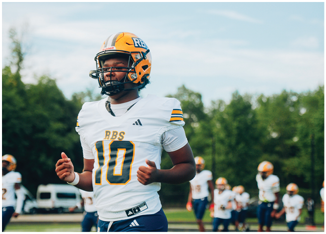 A person wearing a yellow and white football helmet and a white jersey stands outdoors with trees and a blue sky in the background. Description generated by AI