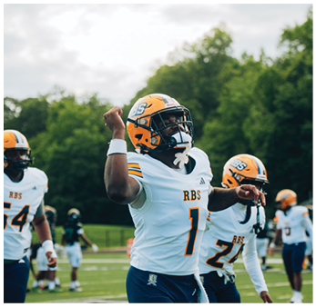A person wearing an orange football helmet is raising a fist outdoors with trees and a cloudy sky in the background. Description generated by AI