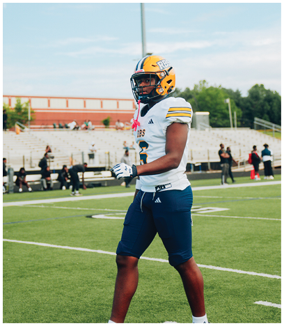 A person wearing a yellow football helmet with blue and white stripes is standing outdoors under a partly cloudy sky with a tall stadium light pole in the background. Description generated by AI