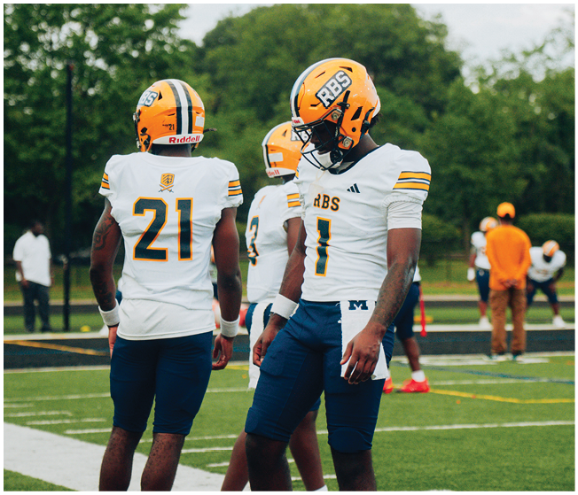 Three people in football uniforms and helmets stand on a field with two others in the background near the sideline. Description generated by AI