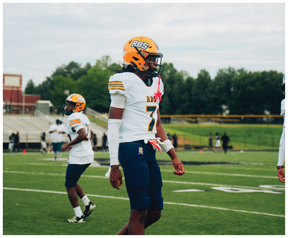Two people wearing football helmets and uniforms stand on a grassy field with bleachers and trees in the background. Description generated by AI