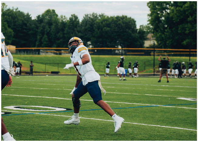 A person in a football uniform is running on a grassy field with other players and a coach visible in the background. Description generated by AI