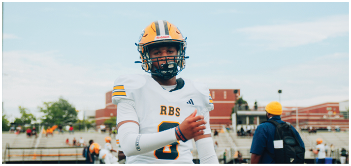 A person wearing a football helmet and white jersey with “RBS" on it stands outdoors on a field with buildings and other people in the background. Description generated by AI