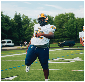 A person wearing a football uniform with a helmet is standing on a field with one leg raised, appearing to stretch or warm up. Description generated by AI