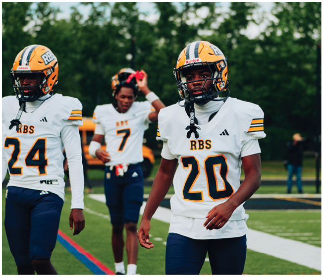 Three people in football uniforms and helmets are walking on a field with trees in the background. Description generated by AI