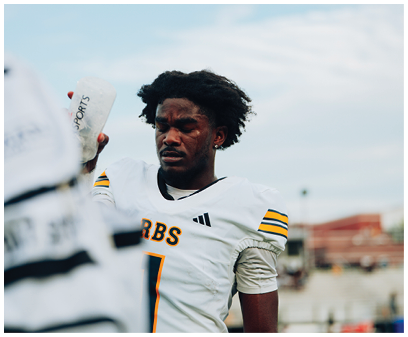 A person wearing a white football jersey with the number one and the letters RBS stands on a field with a focused expression while another person in similar attire holds a water bottle nearby. Description generated by AI