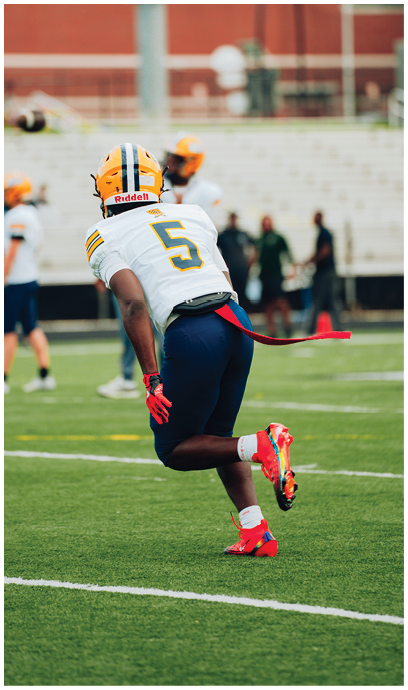 A person wearing a yellow helmet and white jersey with the number 5 is running on a football field with red cleats and gloves. Description generated by AI