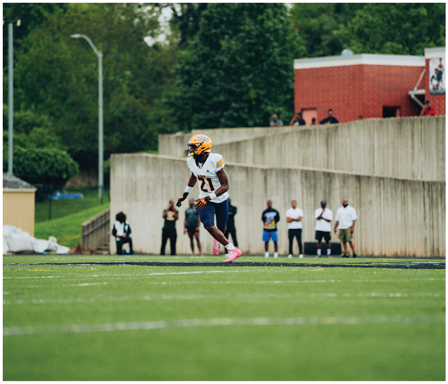 A person wearing a yellow helmet, white jersey, and pink shoes is running on a football field with several people standing in the background near a concrete wall. Description generated by AI