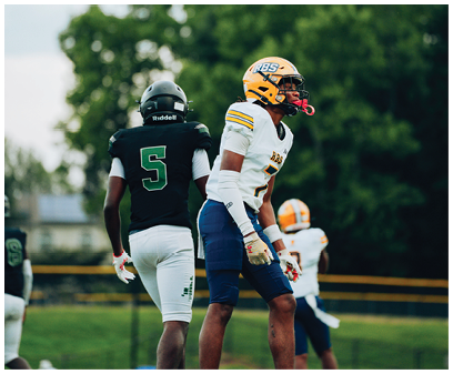 Two people wearing football uniforms and helmets stand on a field with trees in the background, one facing forward and the other facing away. Description generated by AI