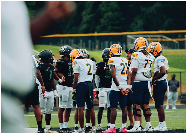 A group of football players in helmets and uniforms are gathered on a field, seemingly preparing for a play or discussing strategy. Description generated by AI
