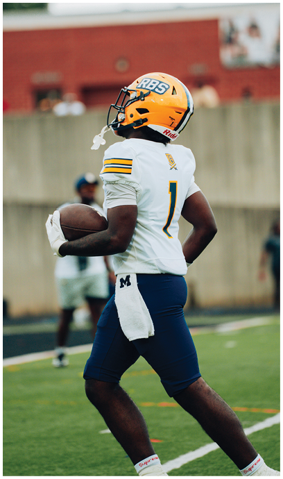 A person wearing a yellow football helmet and white jersey with the number one is holding a football and walking on a grassy field. Description generated by AI