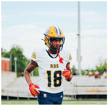 A person wearing a football helmet, white jersey with the number 18 and RBS on it, blue pants, and red gloves is walking on a football field. Description generated by AI