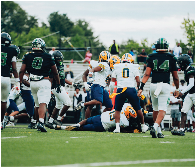 Several people wearing football uniforms are engaged in a play on a grassy field with some players standing and others on the ground. Description generated by AI
