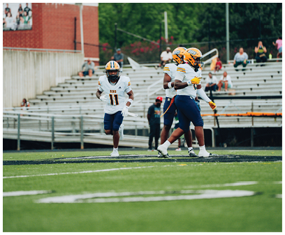Three people wearing football uniforms and helmets are on a football field with empty bleachers in the background. Description generated by AI