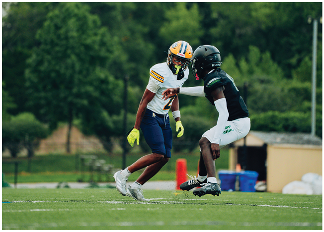 Two people wearing football uniforms and helmets are engaged in a play on a grassy field with trees and equipment in the background. Description generated by AI