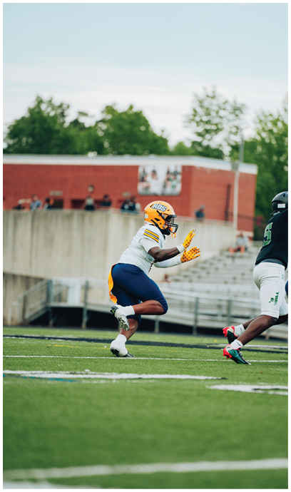 A person wearing a yellow helmet and gloves is running on a football field toward another person in a green and white uniform. Description generated by AI
