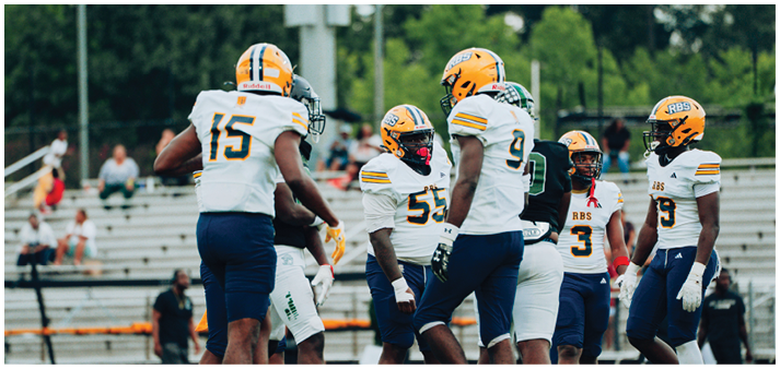 A group of people wearing yellow helmets and white football uniforms are gathered on a field with empty bleachers in the background. Description generated by AI