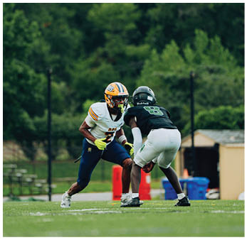Two people wearing football uniforms are facing each other on a grassy field with trees and a small building in the background. Description generated by AI