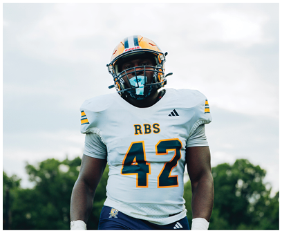 A person wearing a white football jersey with the number 42 and a helmet stands outdoors with trees and a cloudy sky in the background. Description generated by AI