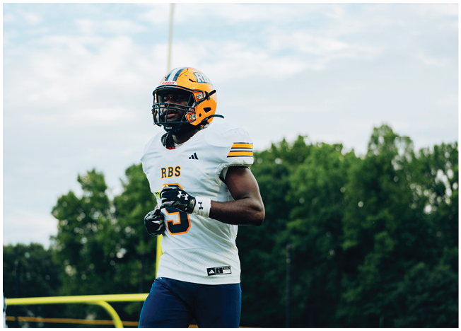 A person wearing a yellow helmet and white football jersey with the letters RBS is standing on a field with trees and a cloudy sky in the background. Description generated by AI