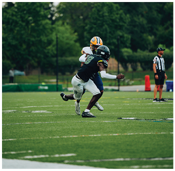 Two people in football uniforms are engaged in a tackle on a green field with a referee standing in the background. Description generated by AI