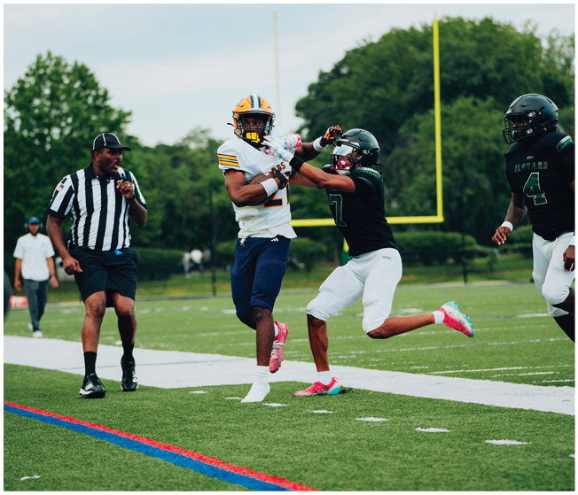 Two people in football uniforms are competing for the ball near the sideline while a referee watches closely on a grassy field. Description generated by AI
