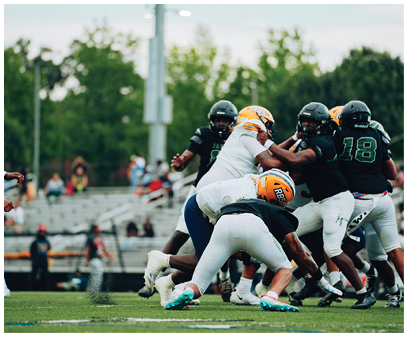 Several people wearing football helmets and uniforms are engaged in a tackle during a game on a grassy field with blurred spectators in the background. Description generated by AI